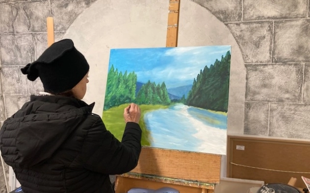 A person wearing a black coat and black hat stands in front of an easel, painting a landscape of a river flowing through a forest under a blue sky. The setting appears to be an indoor studio.