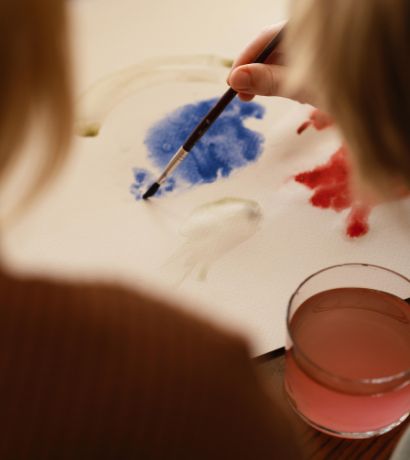 Two individuals are painting on a white canvas. One is holding a brush with blue paint, creating a watercolor effect. There are red and blue paint areas on the canvas. A glass of pinkish water is placed nearby. Both individuals are partially visible.