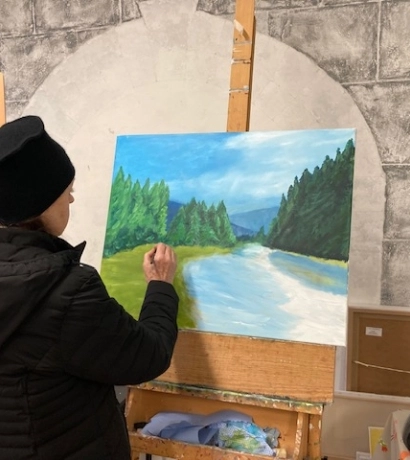 A person in a black jacket and hat paints a landscape on a canvas, featuring a river, trees, and mountains, on an easel indoors. The wall behind has a stone-like arch pattern.