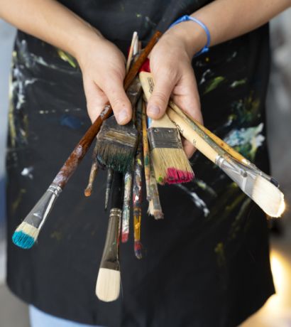 Close-up of a person holding a variety of paintbrushes with painted tips ranging in different sizes and colors. The individual is wearing a black apron with paint splatters on it.