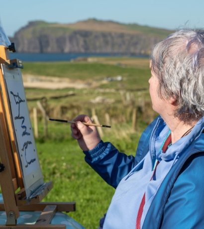 A person with short gray hair paints on an easel outside, overlooking a green landscape with a body of water and cliffs in the background. They are wearing a blue jacket and holding a paintbrush.