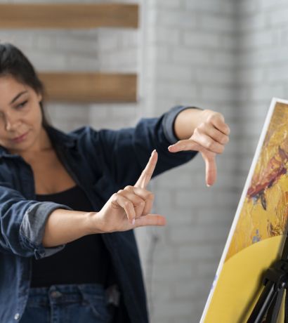 A woman wearing a black top and denim jacket stands near a canvas on an easel, framing her fingers in a rectangle shape as if visualizing the composition. The background features light brick walls and wooden shelves.