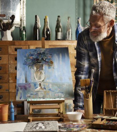 An older adult with gray hair and a beard, wearing glasses and a plaid shirt, is standing in an art studio, looking at a painting on an easel. The painting depicts a vase of flowers. Various art supplies, including brushes and paint tubes, are on the table.