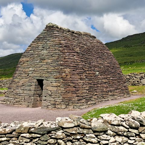 A historic stone structure with a beehive-like shape stands in a grassy landscape against a backdrop of green hills and a partly cloudy sky. The structure is made of stacked stones with a small entrance and a dry stone wall in the foreground.