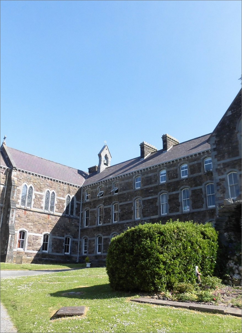 A large, historic stone building with multiple windows and gabled roofs, set against a clear blue sky. The building is surrounded by a grassy area, with a well-manicured bush in the foreground.