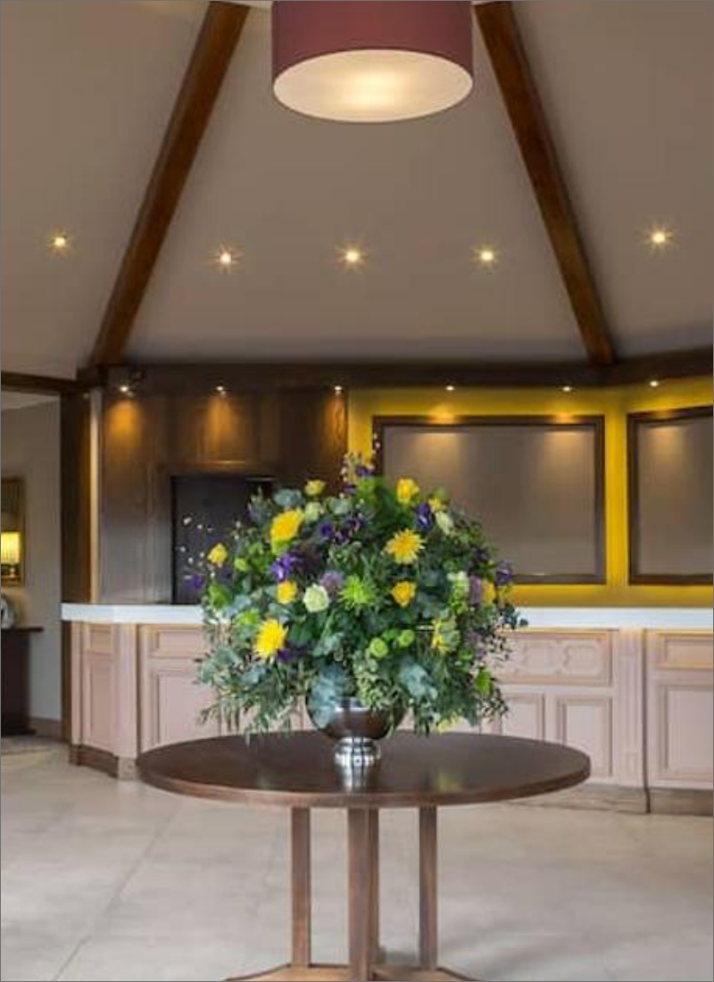 A stylish interior with a round wooden table at the center, adorned with a large, colorful floral arrangement. The room features beige walls, a wooden ceiling with spotlights, and a modern reception desk in the background with yellow and dark brown accents.