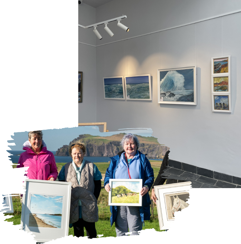 Three people hold paintings in a gallery setting. The gallery wall displays several seascape and landscape artworks. The trio appears to be standing outdoors, with coastal scenery in the background.