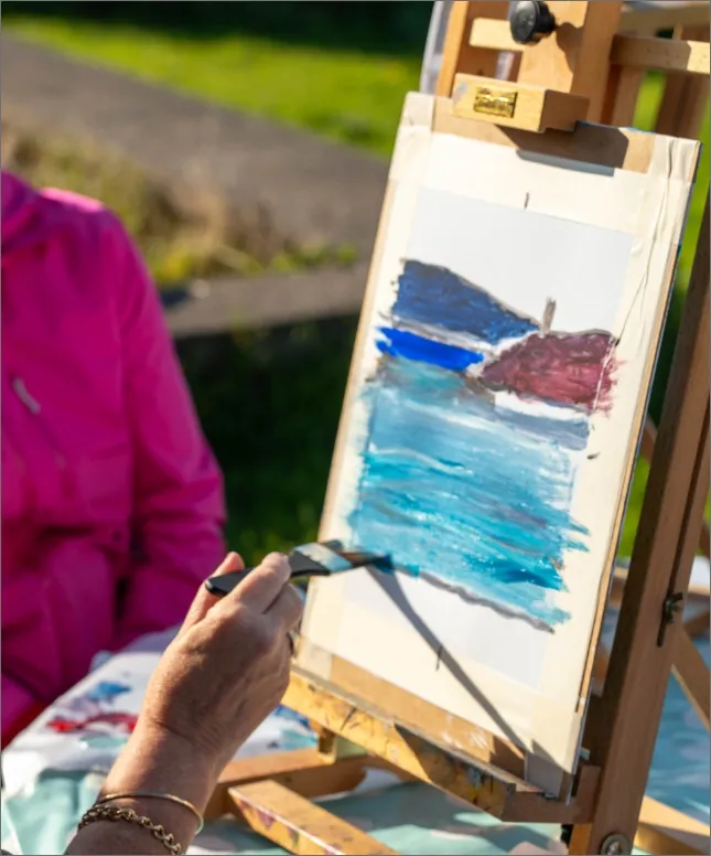 A person in a pink jacket paints a seascape with a brush on a canvas set on an easel outdoors. The scene includes blue water, a hillside, and a small structure.