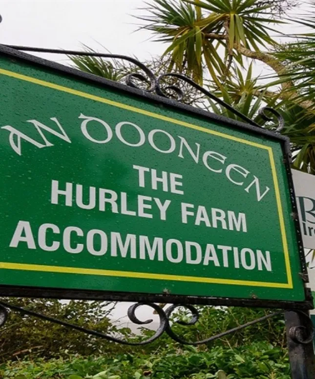 A green sign reads "AN DOONEEN, THE HURLEY FARM ACCOMMODATION" surrounded by lush foliage and a partly visible palm tree.