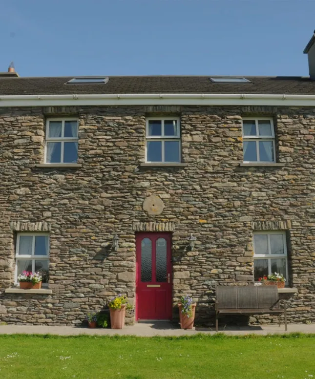 A stone house with a red door, four windows on the upper floor and four on the lower floor. Potted plants with flowers flank the entrance. A wooden bench sits to the right. A grassy lawn is in the foreground under a clear blue sky.