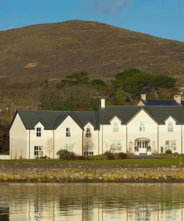 A large white house with a dark roof sits near a calm body of water, surrounded by greenery and backed by a large hill under a clear blue sky.