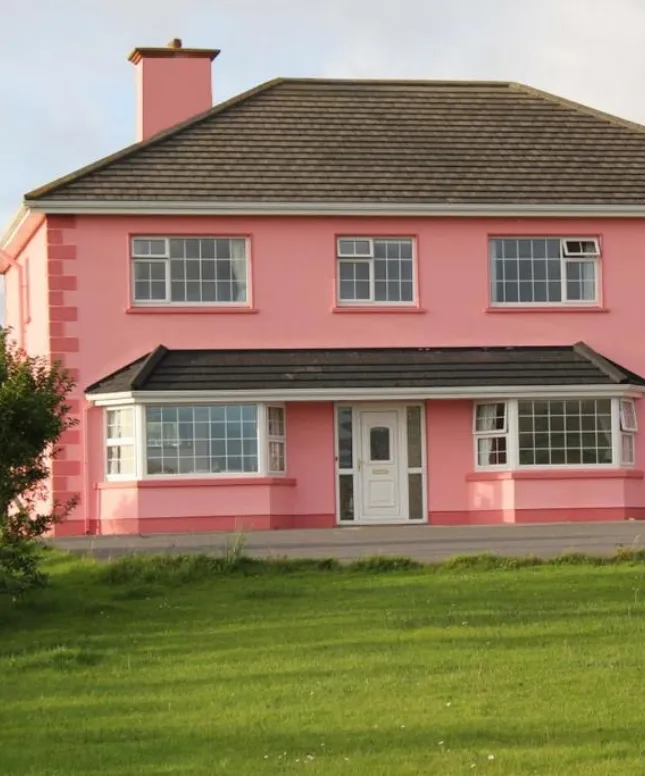 A pink two-story house with a white front door and multiple windows, situated in a grassy area. The sky is clear, suggesting a sunny day. The house has a black roof and a small chimney.