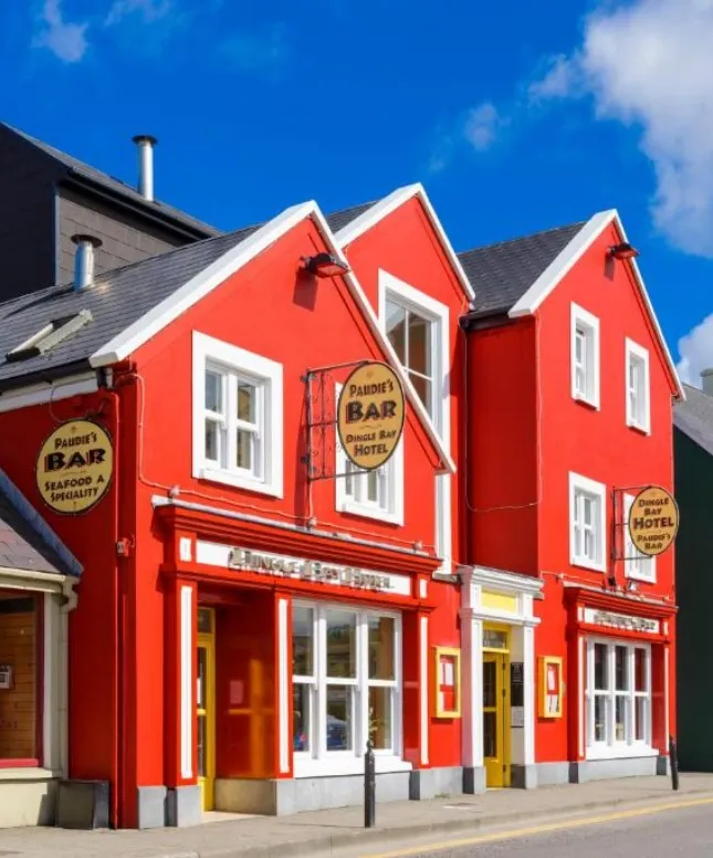 A bright red, two-story building under a blue sky, featuring signs for "Paddy's Bar" and "Dingle Bay Motel." The structure has multiple windows, a sloped roof, and traditional architectural details, situated next to a street.