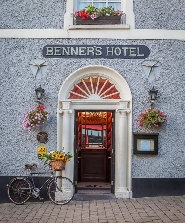 A quaint hotel entrance features a sign reading "Benner's Hotel." The arched doorway is adorned with flowers, lanterns, and a plaque. A vintage bicycle with a basket of sunflowers is parked by the door on the cobblestone pavement.