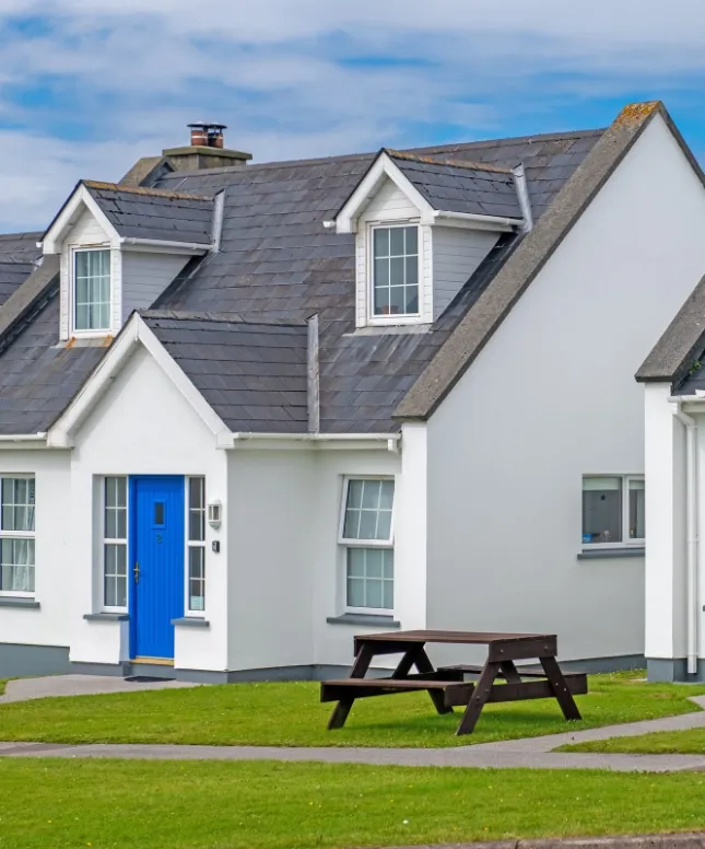 A white, modern cottage with a dark gray slate roof and dormer windows. The house features a bright blue door and a small front yard with a wooden picnic table on the grass. The sky is partly cloudy.