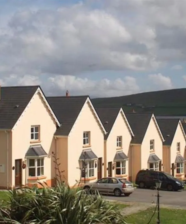 A row of identical cream-colored houses with dark roofs and small front gardens, parked cars in front, set against a backdrop of green hills and a cloudy sky.