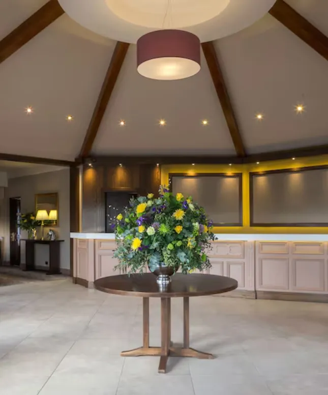 A round wooden table with a large floral arrangement stands in the center of a spacious room. The ceiling has wooden beams and a large hanging light fixture. Soft lighting highlights the modern decor and neutral-toned walls.