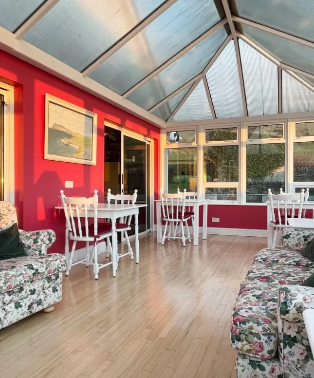 A sunlit conservatory with red walls and a glass roof. The room features floral-patterned sofas and white wooden dining sets. The floor is light wood, and there are artworks on the walls. Sunlight streams in, creating a warm atmosphere.