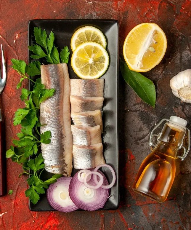 A rectangular black plate with sliced fish, parsley, lemon slices, and red onion rings. The background includes a halved lemon, garlic, a fork, and a small bottle of olive oil on a textured red surface.