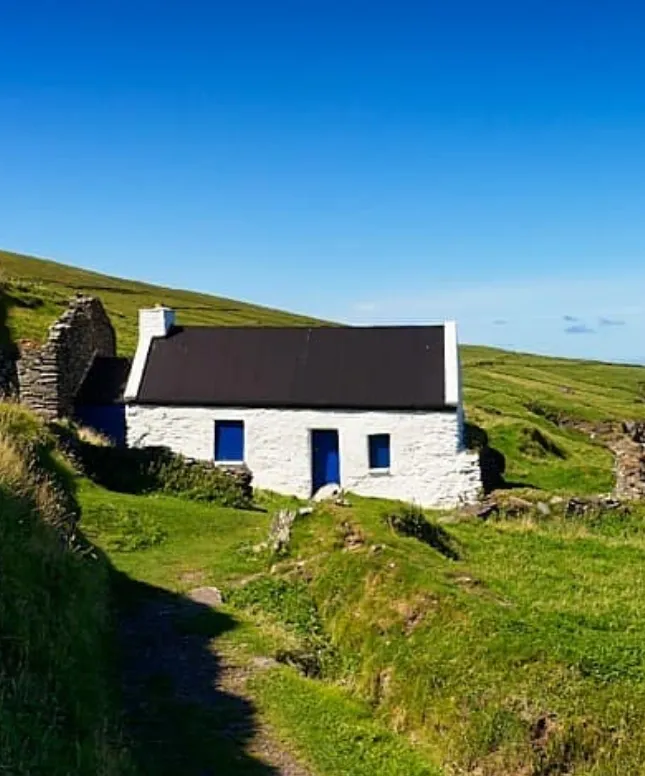 A small white stone cottage with a black roof and blue doors sits peacefully amid lush green hills under a clear blue sky. Ruins of an old stone structure are nearby, adding character to the idyllic rural landscape.