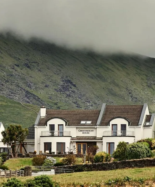 A large house sits at the foot of a misty, green hillside, partially obscured by clouds. The building has a symmetrical facade with arched windows and a well-kept garden in front. A stone wall and fence border the property.