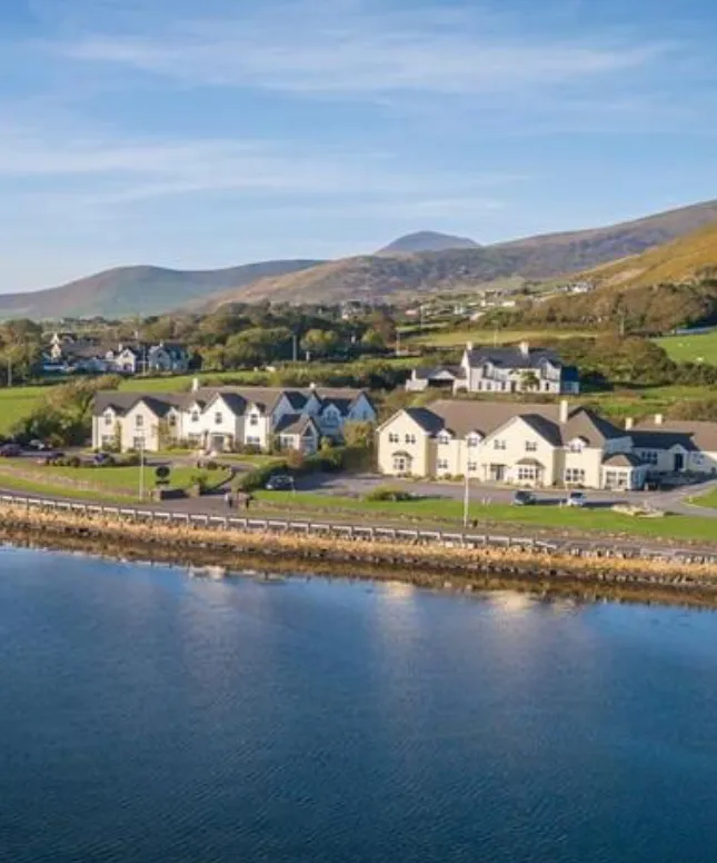 Aerial view of a coastal village with white houses lined along a waterfront road. Green hills and mountains are in the background under a clear blue sky.