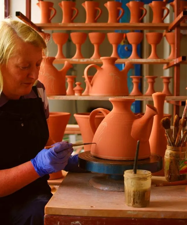 A person wearing blue gloves is painting a large, unfinished clay teapot on a spinning wheel. In the background, shelves are filled with similar pottery pieces. Paintbrushes and a pot of paint rest nearby on the table.