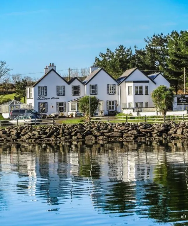 The image shows a white building labeled "Milltown House" situated by a calm body of water. The water reflects the house, surrounded by trees and greenery. The sky is clear and blue, enhancing the serene atmosphere.