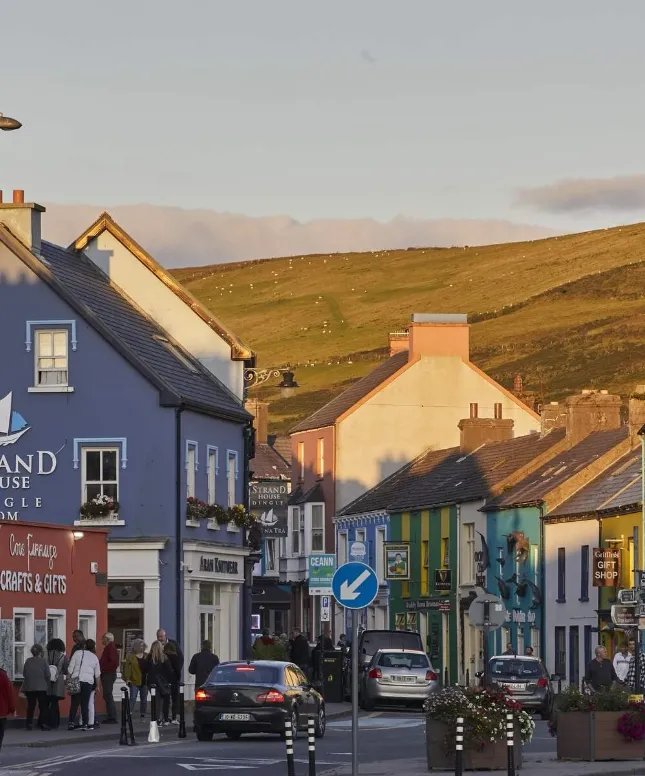 A vibrant street scene in a small town features colorful buildings under a clear sky. People walk on the sidewalk, and distant hills covered in greenery are visible in the background.