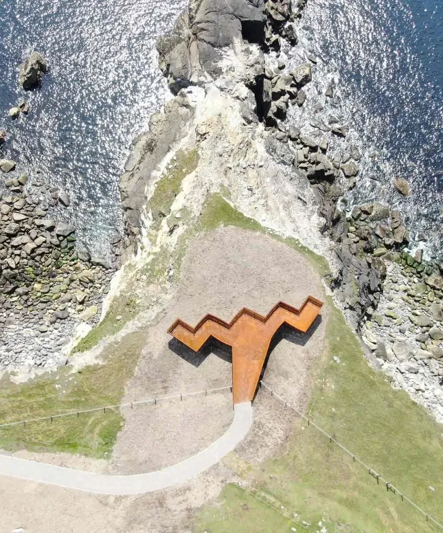 Aerial view of a zigzag-shaped rusty metal sculpture on a rocky coastline. The sculpture sits on grass near the edge of a cliff, with the blue ocean and rocky outcrops below. A winding path leads to the sculpture.