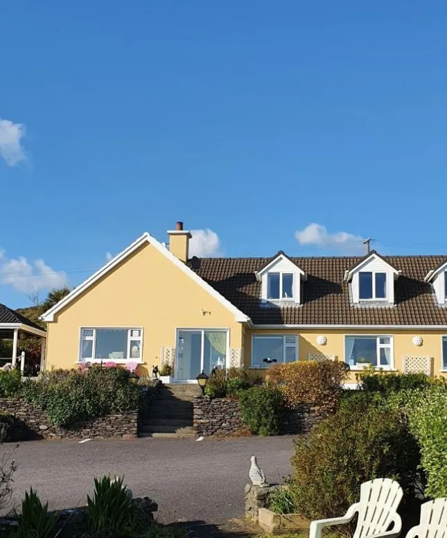 A charming yellow house with a sloped roof and white-framed attic windows set against a clear blue sky. The front yard features a stone wall, shrubs, and a white chair. A small statue sits in the garden to the right.