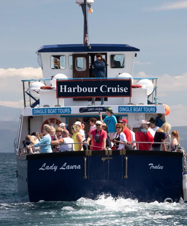 A boat named "Lady Laura" with "Harbour Cruise" and "Dingle Boat Tours" signs. It's filled with people enjoying a sunny day on the water. The ocean is calm, and the sky is clear with a few clouds.