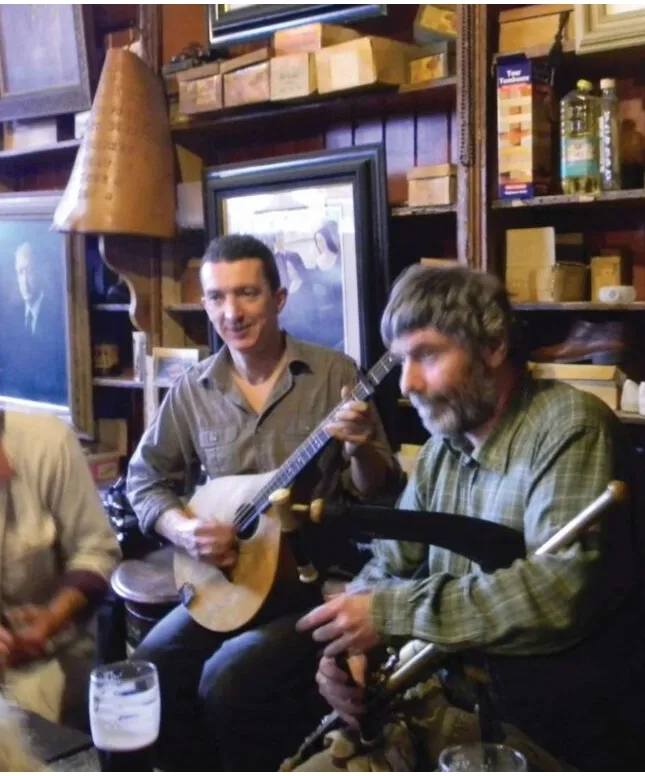 Two musicians are playing instruments in a cozy room filled with framed pictures and wooden shelves. One plays a string instrument, while the other plays a bagpipe. A third person is partially visible. A glass of beer is on the table.