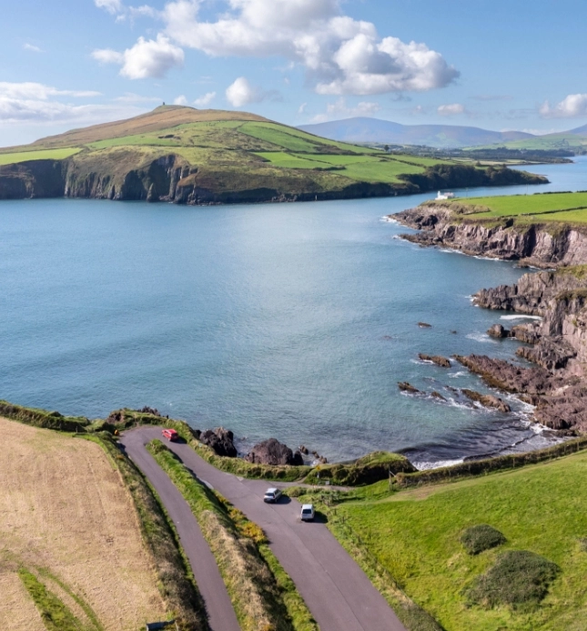 A scenic coastal view features a winding road next to rocky cliffs by a calm blue sea. Green hills and fields roll into the distance under a partly cloudy sky. Three cars are parked along the road.