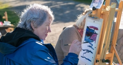 An older adult with gray hair is painting a colorful abstract design on a canvas outdoors, wearing a blue jacket. Another person sits nearby at a wooden easel. Sunlight casts shadows on the ground.