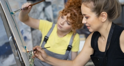 Two women painting on a large canvas. The woman in the foreground has light hair in a bun and wears a black top and apron. The woman in the background has curly red hair and wears a yellow shirt and gray overalls. Both are focused on their work.