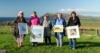 Five people standing on grass holding framed artwork, with an ocean and rocky cliffs in the background under a clear blue sky.