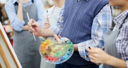 A group of people in aprons participate in a painting class. One person holds a paintbrush and a colorful palette, standing in front of an easel. Others observe and engage, with soft focus on their faces.