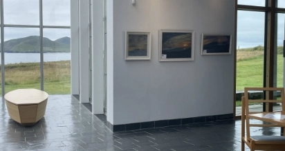 A modern gallery interior with three landscape artworks on the wall. Large windows offer a view of grassy fields and hills by the sea. A wooden bench and part of a chair are visible on the tiled floor.