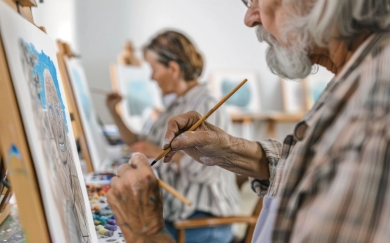 Older adults focus on painting on canvases in a classroom setting. They use paintbrushes and palettes filled with various colors. The room is filled with natural light, enhancing the creative atmosphere.