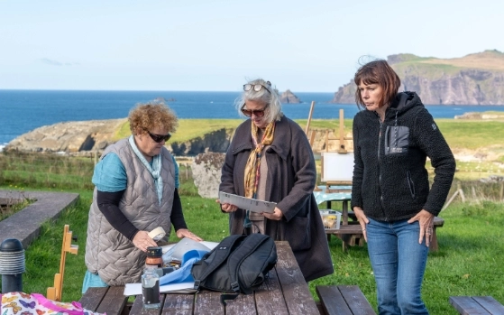 Three women stand around a picnic table on a coastal landscape. Two are looking at an open folder on the table, while the third looks on. The ocean and cliffs are visible in the background under a clear sky.