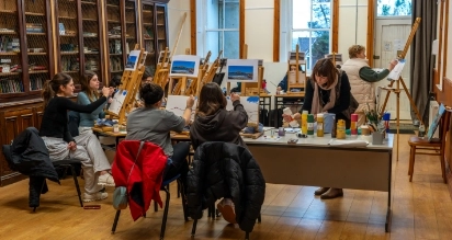 A group of people seated around tables with art supplies, painting landscapes on canvases in a library-like room with bookshelves. An instructor stands at an easel, demonstrating. Coats are draped over chairs, and the room has large windows.