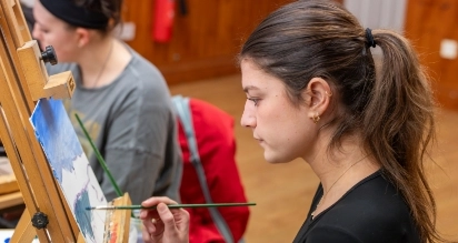 A young woman with long hair in a ponytail concentrates on painting at an easel. She holds a brush, adding details to a landscape on the canvas. Another person is partially visible in the background, also focused on painting.