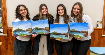 Four people smile while holding their landscape paintings. Each artwork depicts a similar scene of hills and a body of water under a blue sky. They stand indoors against a wooden wall, wearing casual clothing.