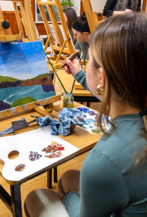 A person sits at a table painting a landscape, featuring hills and a lake, on a canvas. An easel holds the canvas, and a palette with paints is nearby. Other people painting can be seen in the background.