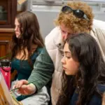 An older woman guides a young woman painting at an easel. The older woman is wearing a light vest and sunglasses atop her curly hair. The younger woman has long dark hair and appears focused. Another young woman paints in the background.