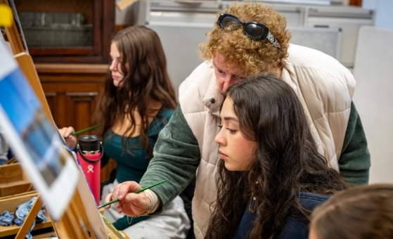 An older woman guides a young woman painting at an easel. The older woman is wearing a light vest and sunglasses atop her curly hair. The younger woman has long dark hair and appears focused. Another young woman paints in the background.