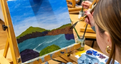 A person paints a landscape on a canvas, depicting green hills and a blue sky. The canvas rests on an easel, and various paints and brushes are visible on the table. The person's hand holds a brush poised over the canvas.