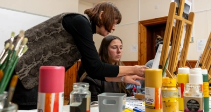 A woman leans over to help another woman who is seated and working at an easel in an art class. Art supplies like paintbrushes and jars of paint are on the table. Both women are focused on the project.