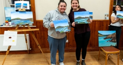 Two people smiling and holding landscape paintings in a room with wood-paneled walls. An easel with more paintings and a chair are visible. A third person is partially seen on the right.